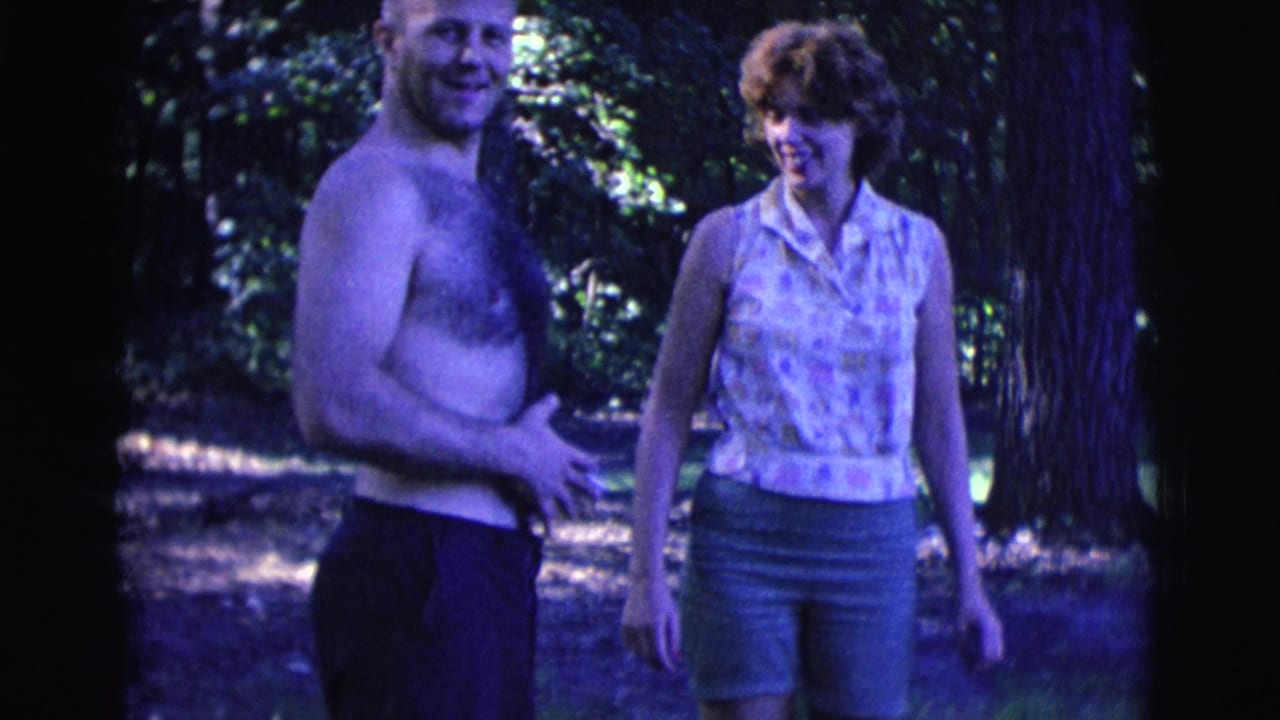 Shirtless man with cigarette stands beside smiling woman in a wooded area.