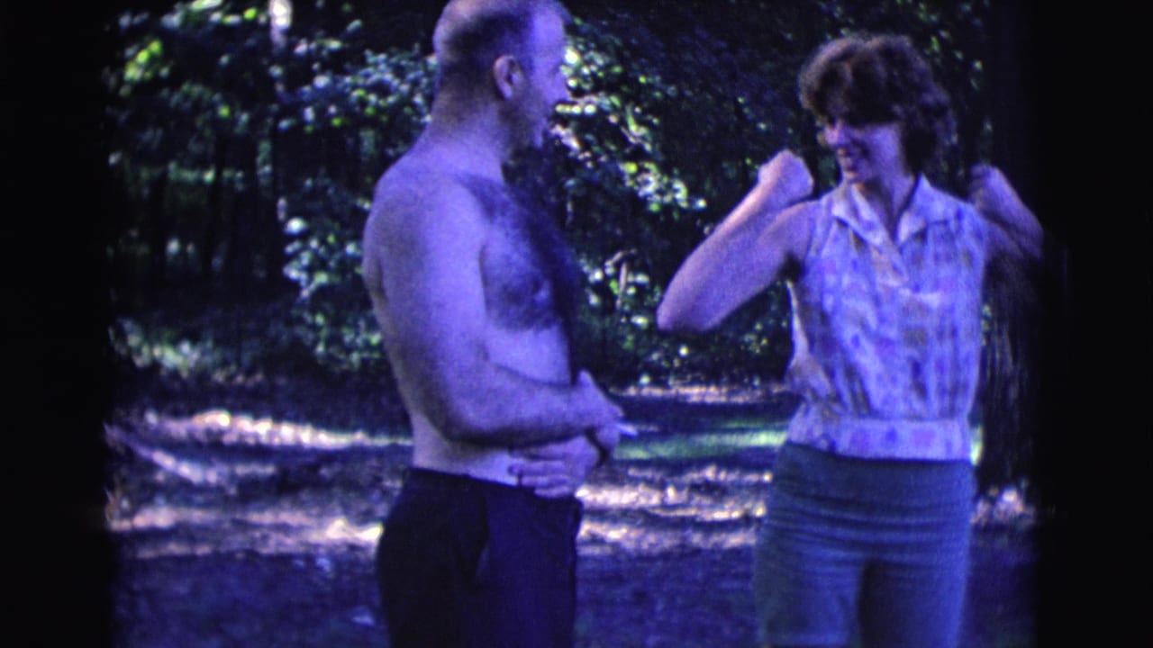 The woman in a sleeveless patterned shirt playfully lifts her arms toward the shirtless man during the Clarkdale 1961 outdoor home movie.
