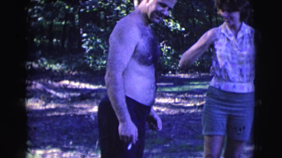 A shirtless man with a cigarette lowers his gaze while the woman reaches toward him in the wooded Clarkdale 1961 home-movie scene.