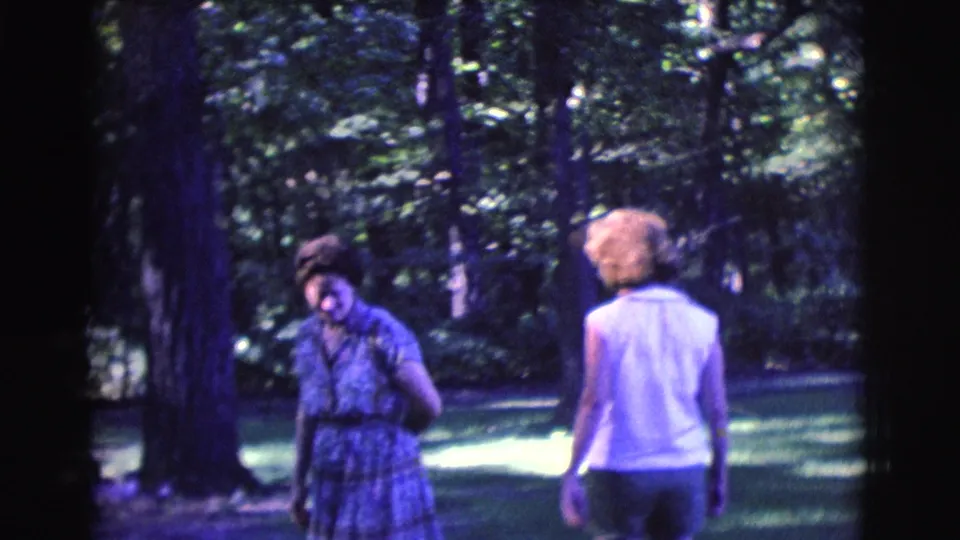 Two women move through a shaded garden, one facing down near a tree and one seen from behind, in 1961 Clarkdale footage.