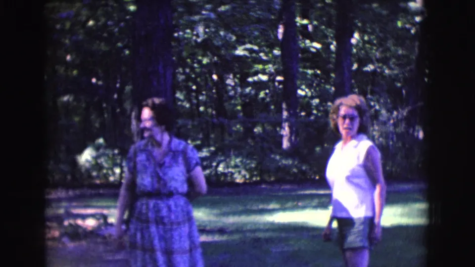 Two adult women walk through a sun-dappled garden in Clarkdale, Arizona, with trees and grass behind them.