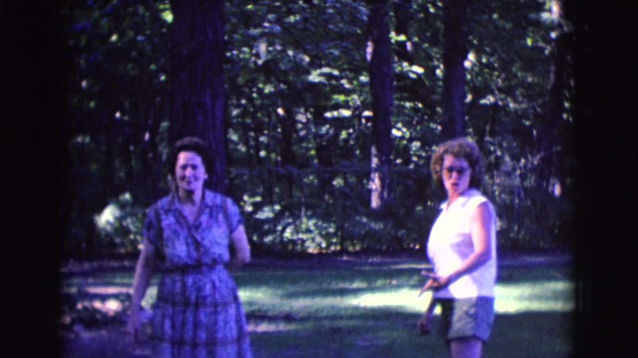 Two adult women stand apart on a shaded lawn with trees behind them.