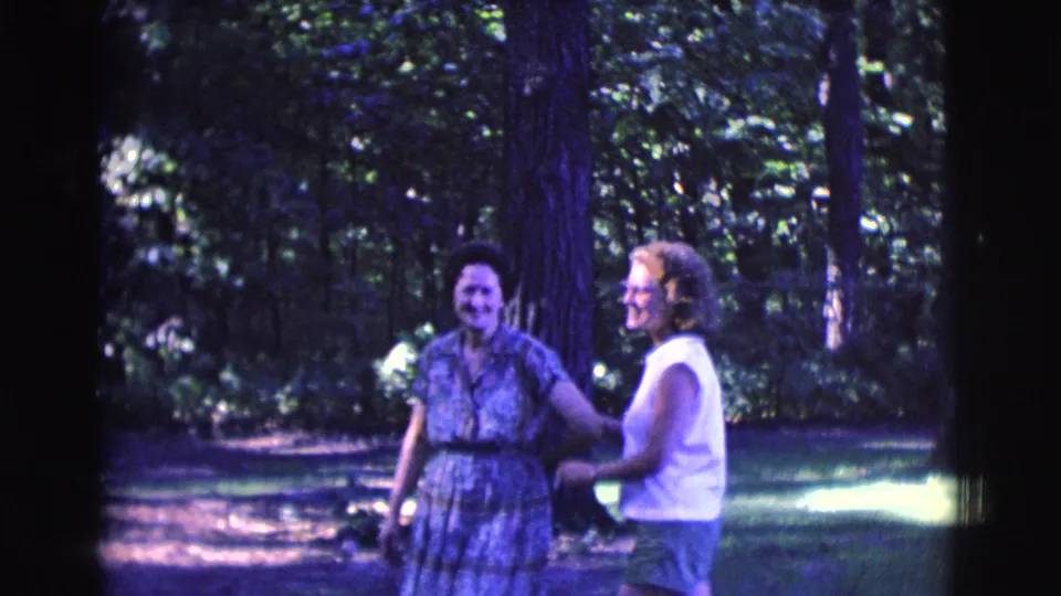 In the 1961 home-movie frame, the women smile and move closer on the shaded grass, suggesting a friendly greeting.