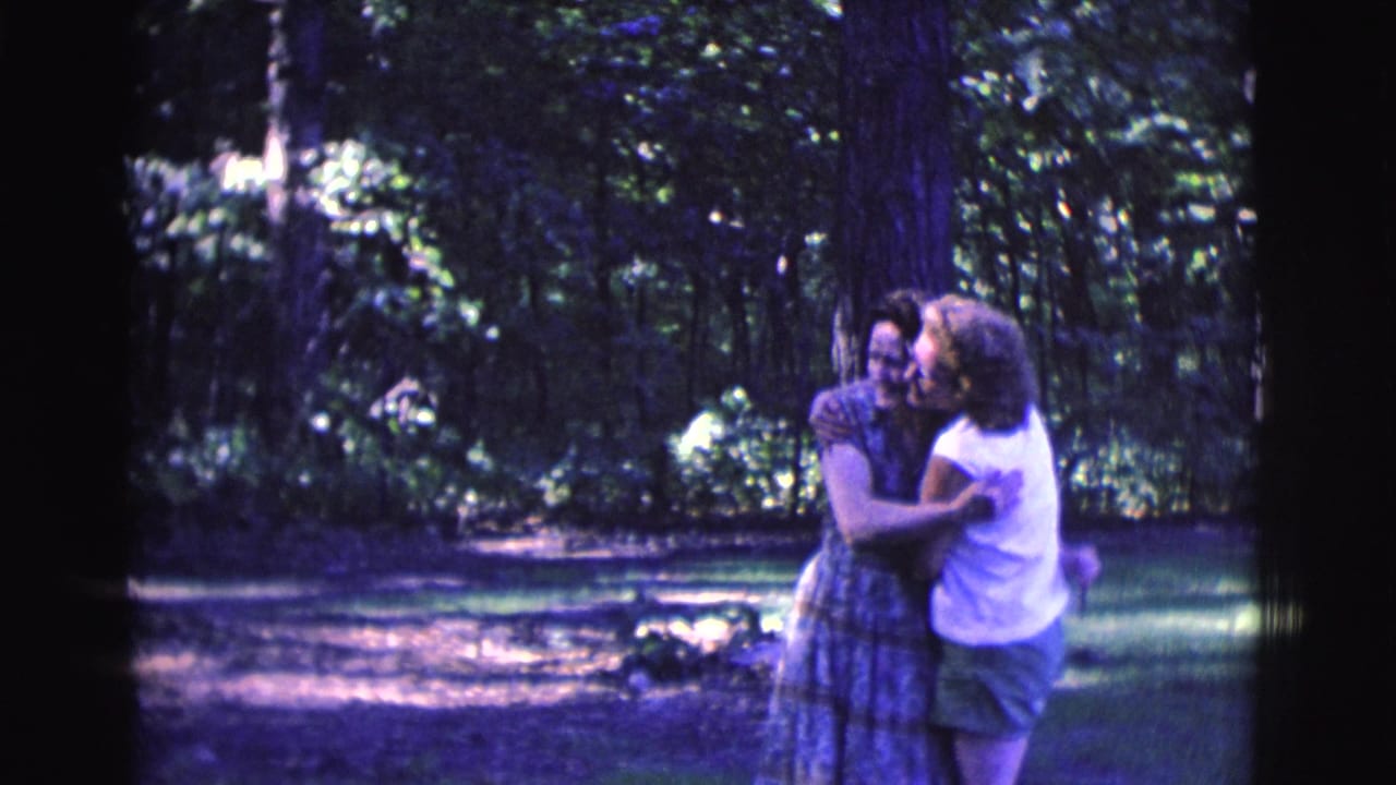 Two adult women embrace in a shaded wooded area while a third stands close beside them.