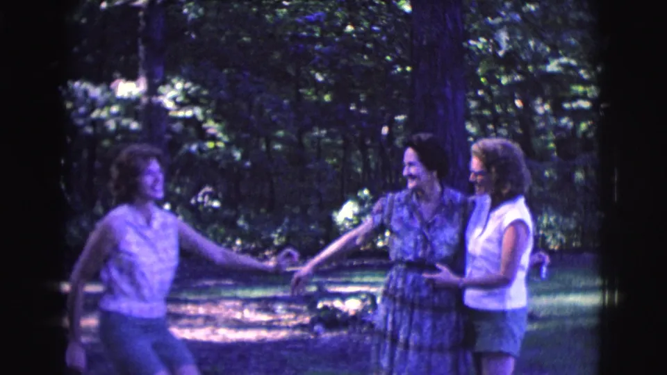 Three adult women in 1961 Clarkdale home-movie footage hold hands playfully in a wooded clearing.