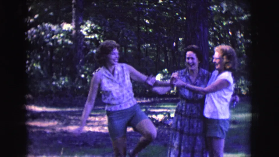 In the wooded Clarkdale scene, three adult women laugh and hold hands while one strikes a playful dancing pose.