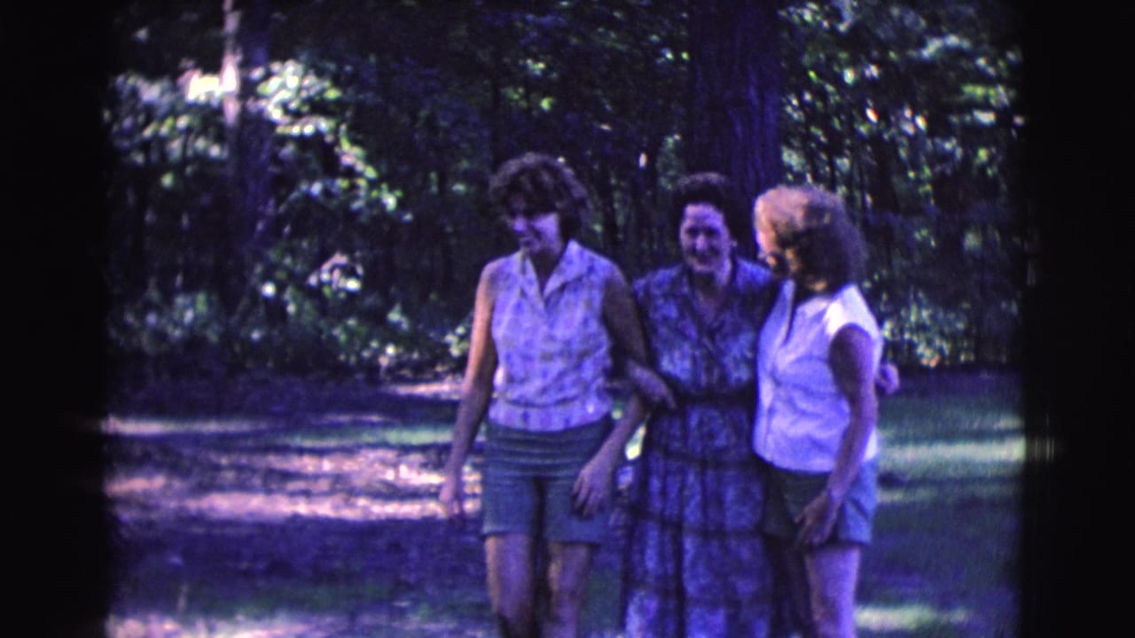 Three adult women walk arm in arm through a wooded outdoor area.