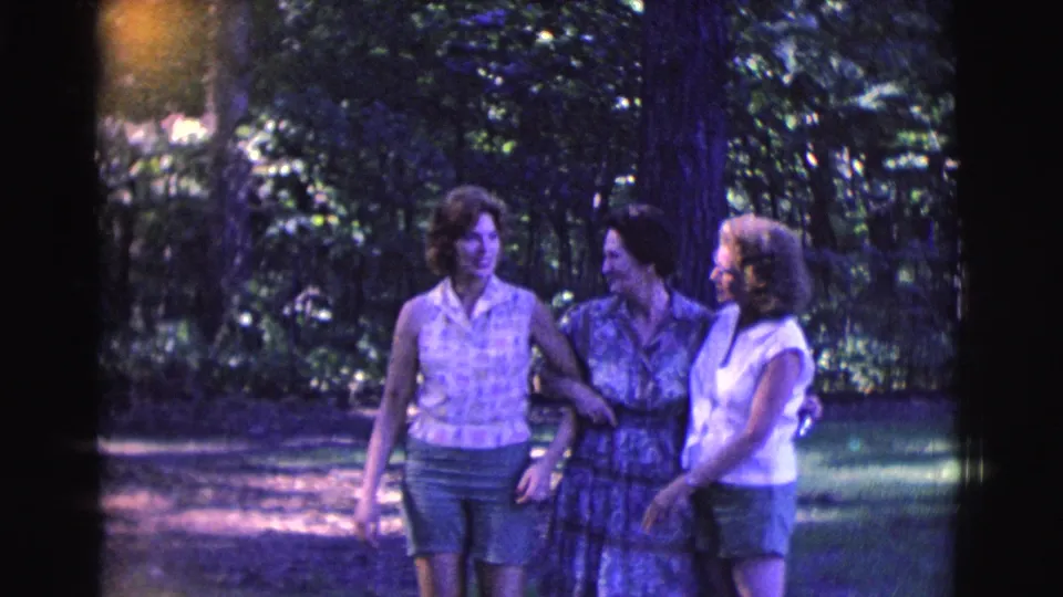 Three women in casual 1961 clothing walk arm in arm through a green, tree-lined outdoor space.