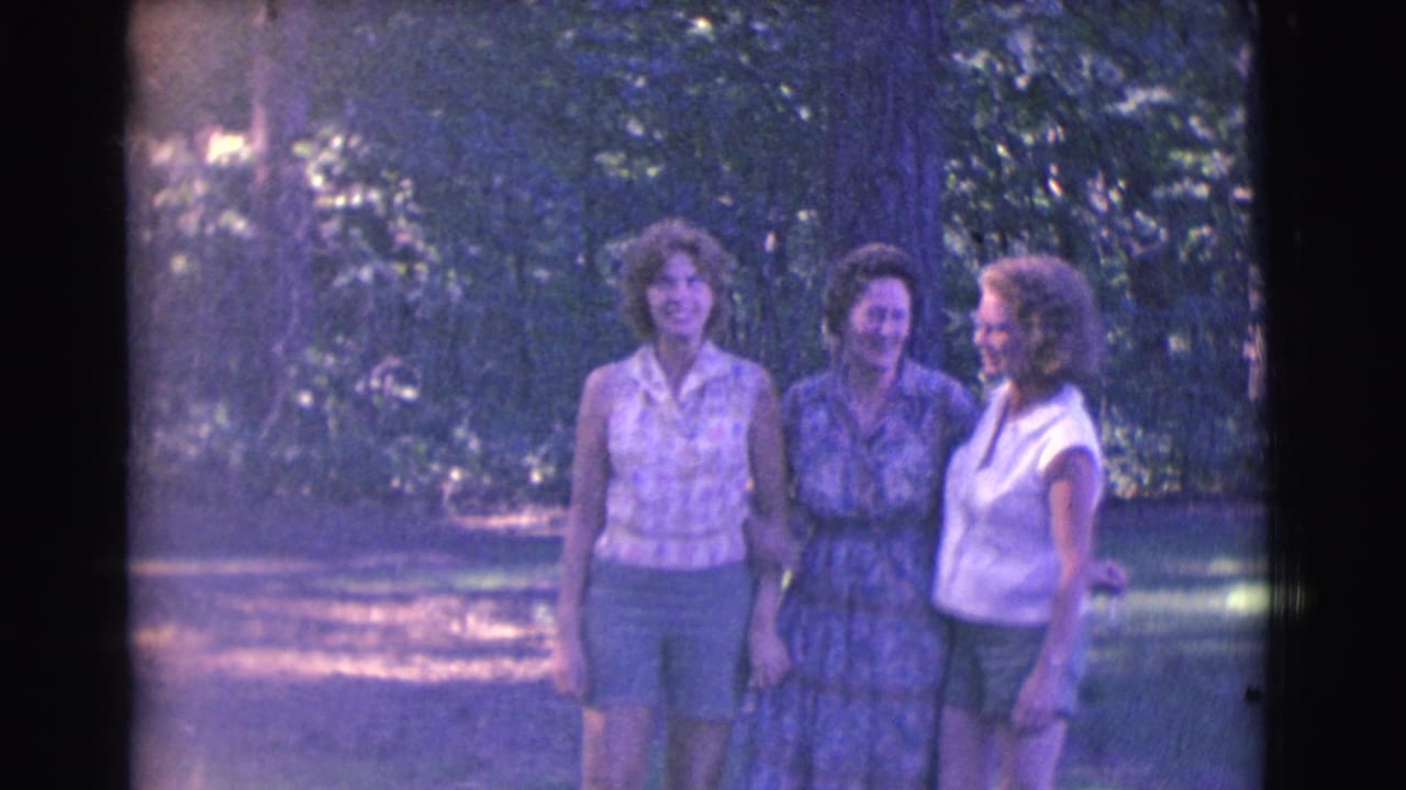 Three adult women stand close together in a wooded area, two facing the camera and one turned in profile.