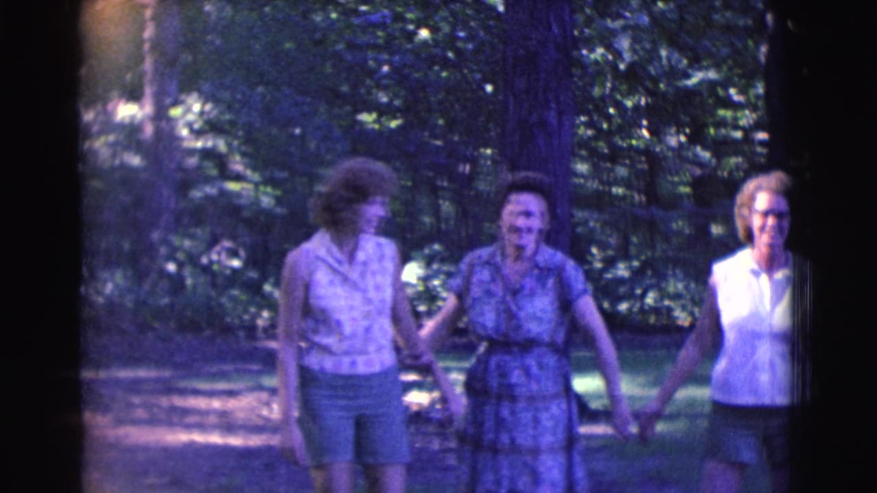 Three adult women walk arm-in-arm through trees, smiling as they move along a shaded path.
