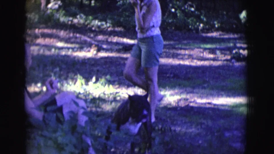1961 Clarkdale, Arizona home-movie frame showing two adults and a black-and-white dog in a sun-dappled wooded area.