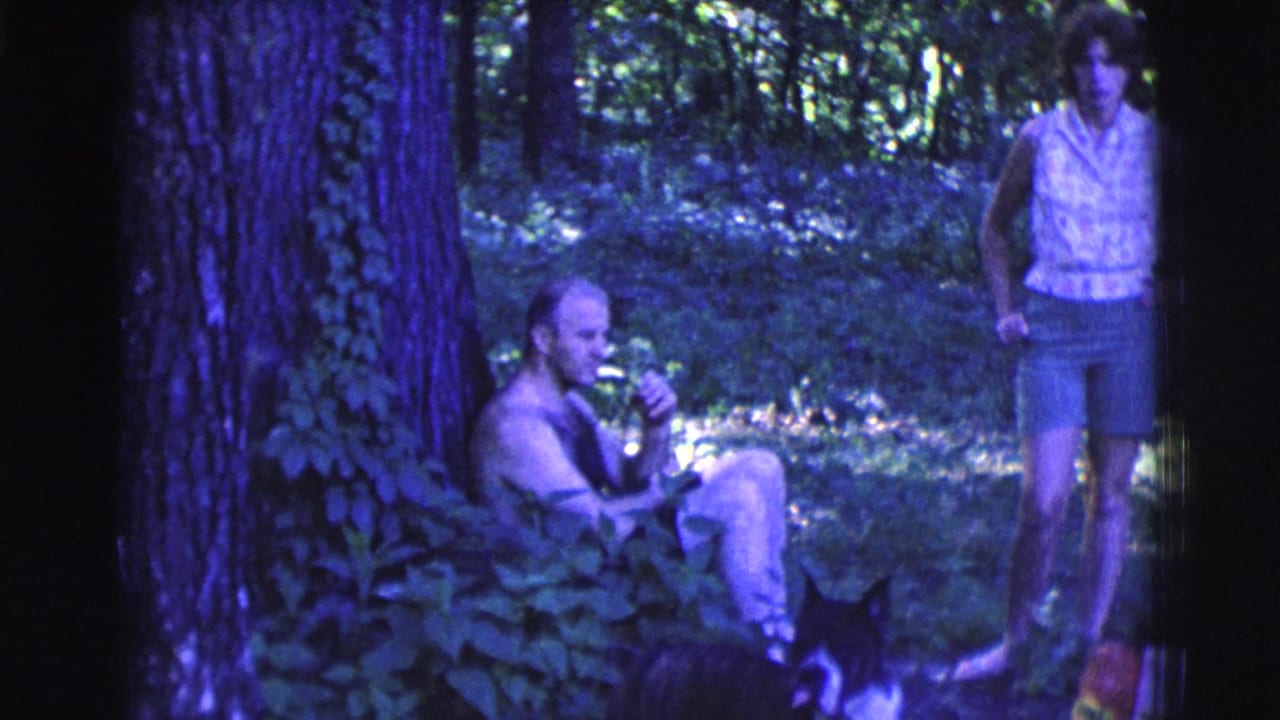 1961 Clarkdale, Arizona frame with a seated man smoking near a tree, a standing woman and a black-and-white dog in woods.