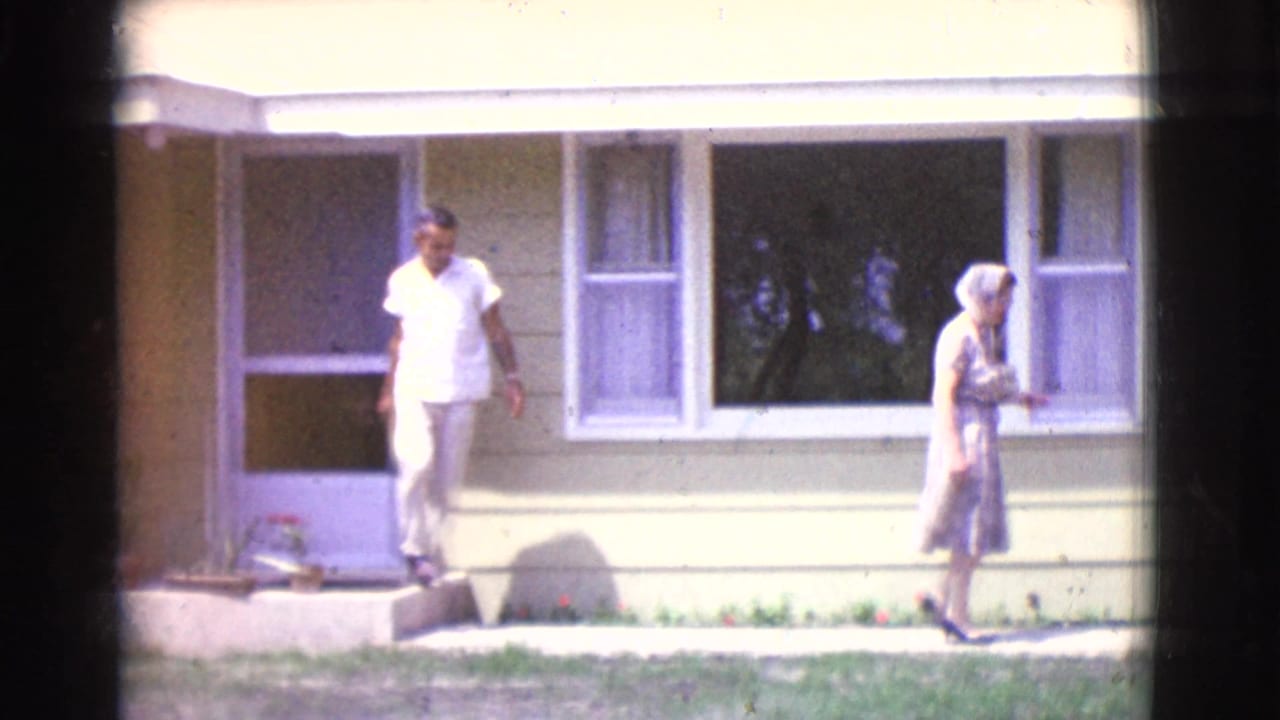 Man steps from a light-colored house while a woman in a dress walks along the sidewalk.