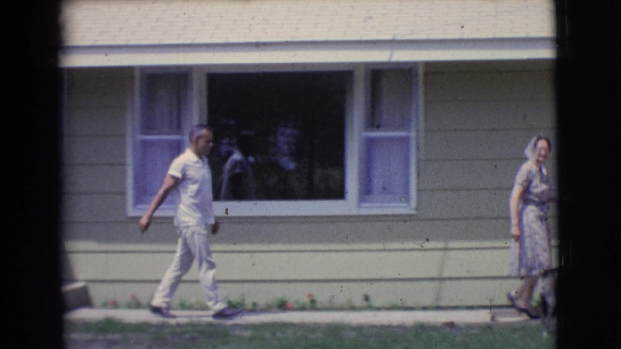 The adults continue along the sidewalk beside the house, with broad windows and horizontal siding visible behind them.