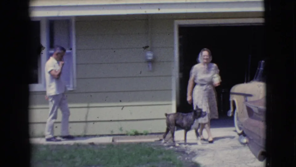 Near the driveway, the woman pauses with a small dog while the man walks by the house exterior and open garage area.