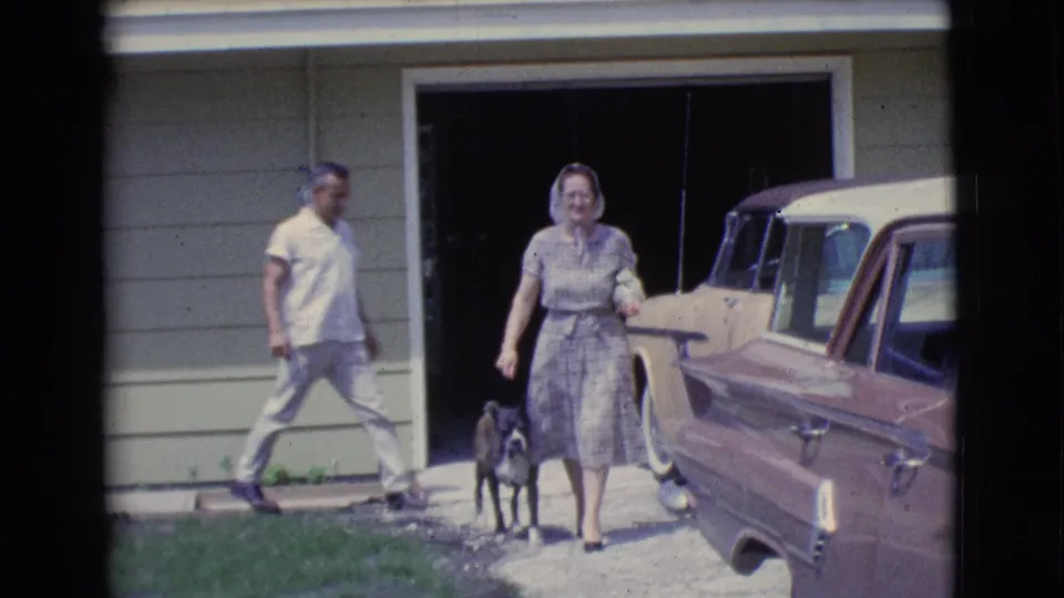 The woman and small dog approach from the driveway area as the man walks past and a parked vintage car fills the right side of the frame.