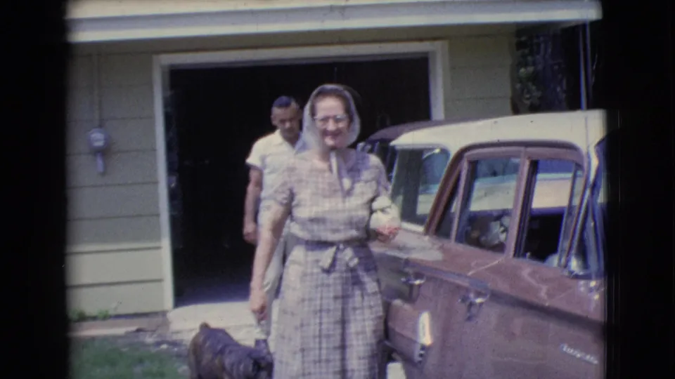 The woman smiles near the brown car, with the man and dog behind her by the open garage in this 1961 Clarkdale home-movie frame.