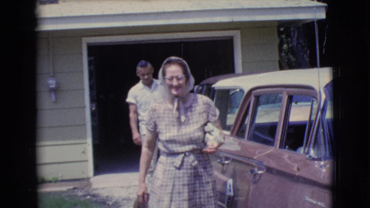 Woman in a checkered dress walks beside a vintage car as a man stands near an open garage.