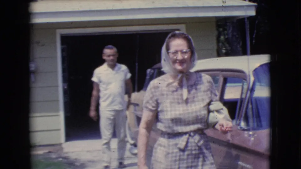 The woman in a headscarf and eyeglasses smiles as she nears the camera, with a man walking behind her from the house garage in Clarkdale footage.