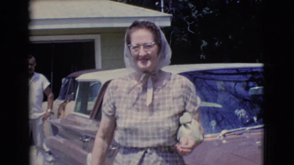 A closer 8mm frame shows the smiling woman in a checkered dress and headscarf walking past a vintage car, with the house and man partly visible behind her.
