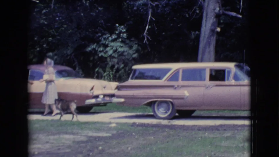 1961 Clarkdale, Arizona home-movie frame showing a woman near a small dog and two parked two-tone cars by trees.