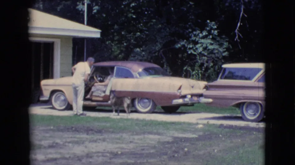 The man remains by the open sedan door in the Clarkdale driveway while the dog stands near the rear of the car and another vehicle is parked nearby.