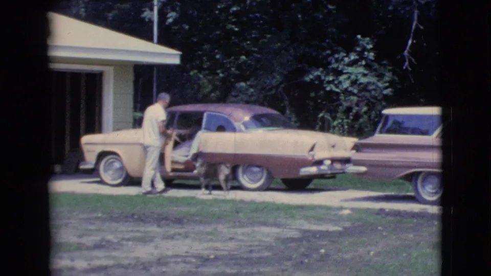 1961 Clarkdale, Arizona home-movie frame showing a man beside a vintage two-tone car, a seated passenger, and a small dog in a driveway.