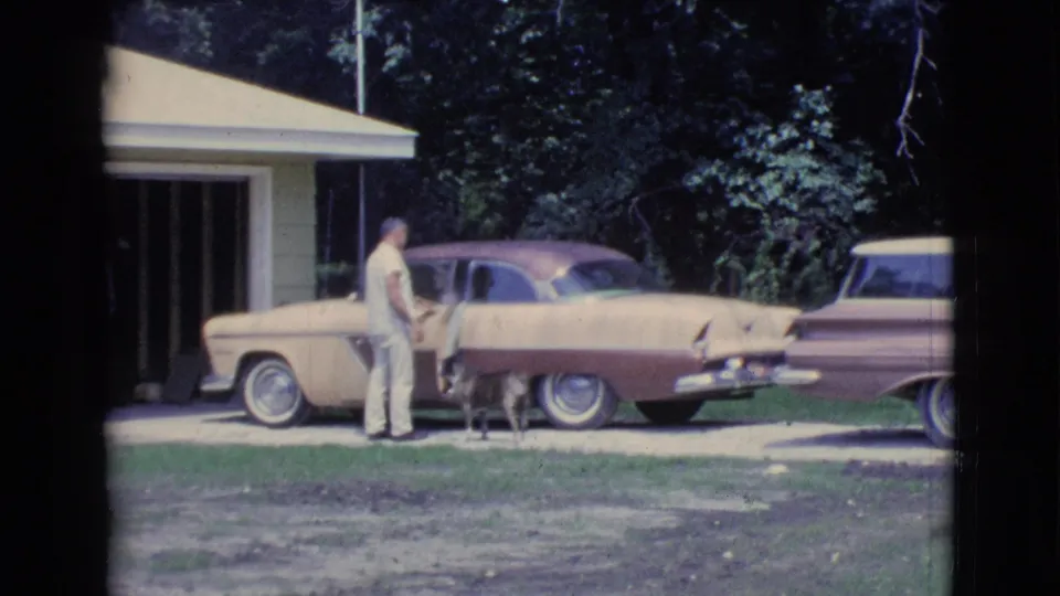 1961 Clarkdale home-movie frame showing a driveway exchange beside a vintage two-tone car, with dog, house, and trees visible.