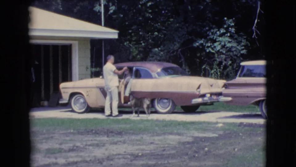 1961 Clarkdale driveway view of a man talking toward someone inside a two-tone parked car as a dog stands close.