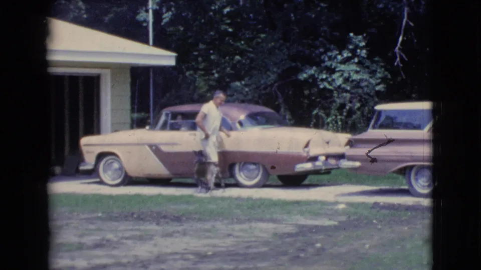 Late frame from the 1961 Clarkdale home movie with a man and dog beside a two-tone car in a driveway.