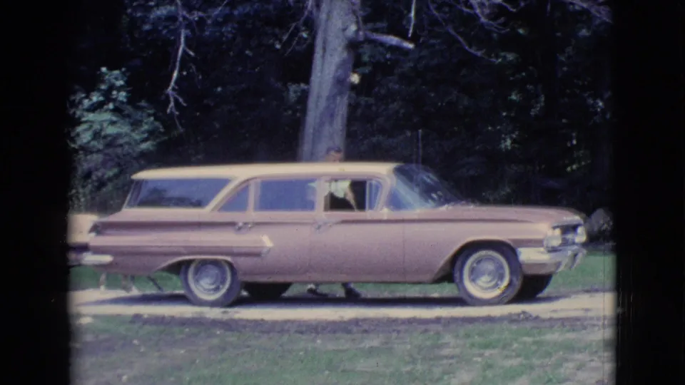 The 1961 home-movie frame shows a side view of the two-tone station wagon parked along the driveway with trees behind it.