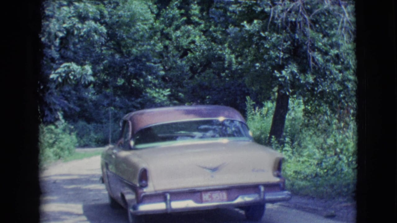 Cream and maroon vintage sedan driving away on a shaded wooded road.