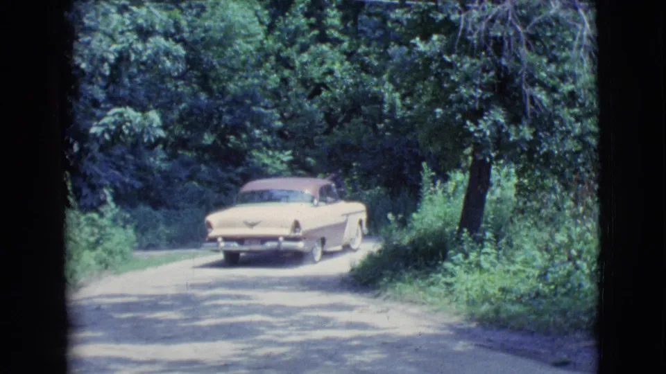 The cream and maroon sedan moves around a wooded bend, with soft 8mm color and grain consistent with the supplied 1961 home-movie source.