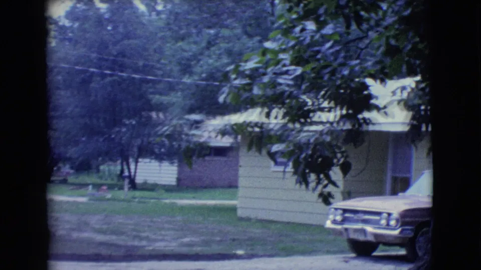 1961 Clarkdale, Arizona frame showing the side of the yellow house, shaded lawn, overhanging leaves, and the front of the parked station wagon.