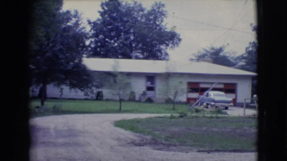 1961 Clarkdale, Arizona frame of a house exterior with a red garage door, parked sedan, curved driveway, grass lawn, and mature trees.