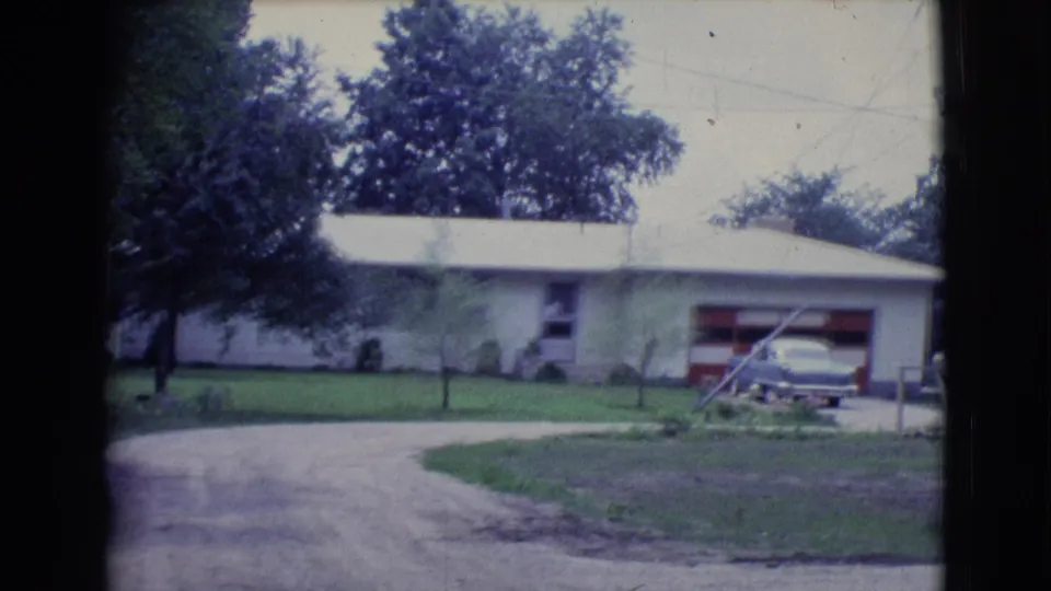 1961 Clarkdale, Arizona home-movie view of the residence with red garage door, older sedan, driveway curve, lawn, and leafy trees.