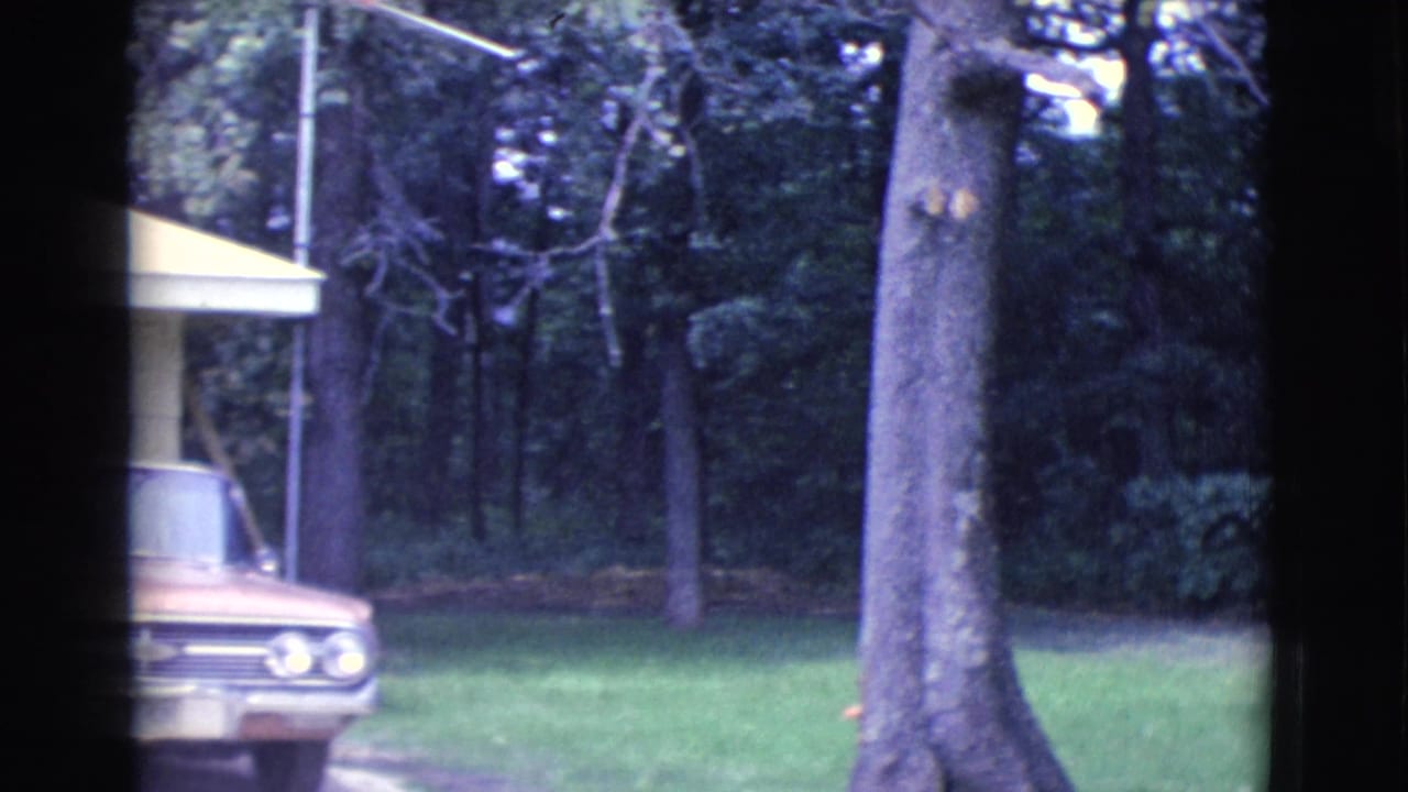 1961 Clarkdale home-movie frame showing a partial yellow ranch house exterior with a parked sedan, green lawn, and mature trees.