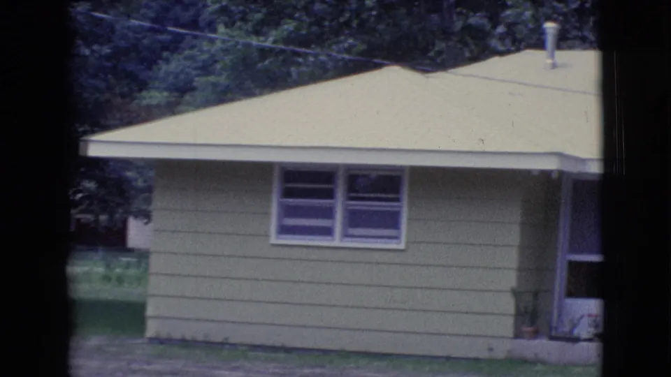 1961 Clarkdale, Arizona frame showing the right side of a light green house, small windows, doorway edge, lawn, and shaded trees.