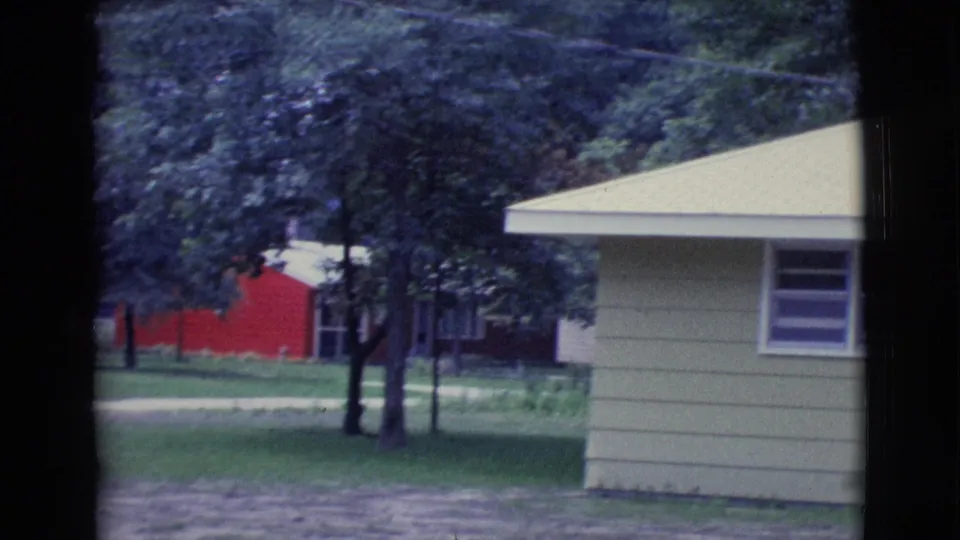 1961 Clarkdale, Arizona 8mm frame with the house edge at right, leafy trees, green yard, and a red building beyond.