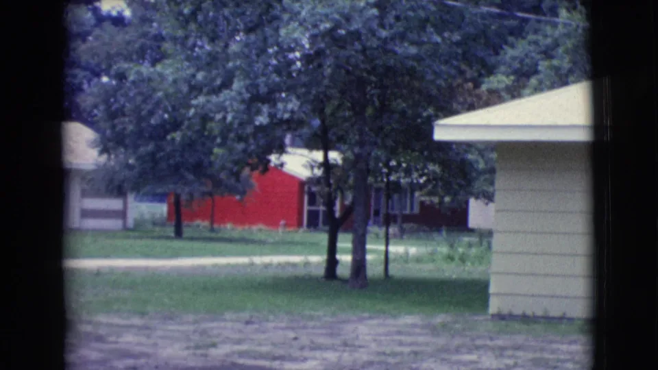1961 Clarkdale, Arizona frame showing the right edge of the house, trees in the yard, green grass, and a red building in back.