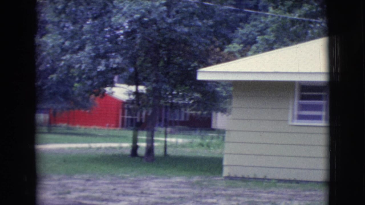 Light green house side with pale roof, trees, lawn, and red building in background.