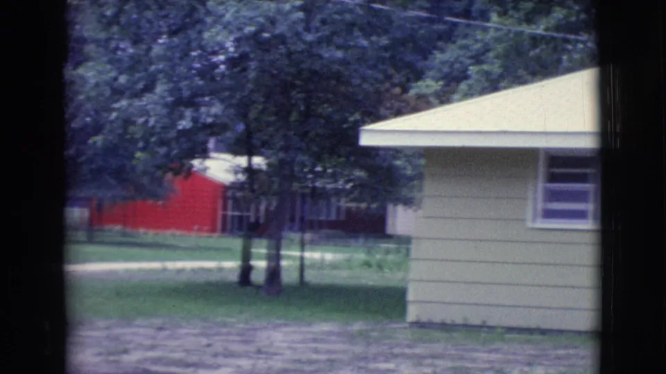 1961 Clarkdale, Arizona 8mm frame showing the side of a light green house with trees, lawn, and a red building beyond.