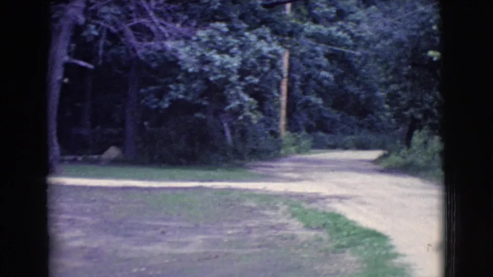 1961 Clarkdale, Arizona frame showing a quiet dirt or gravel road bordered by lawn, trees, and dense greenery.