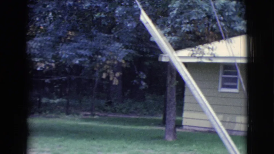 1961 Clarkdale, Arizona 8mm frame showing more of the lawn and trees, with the house edge and a long white pole in the foreground.