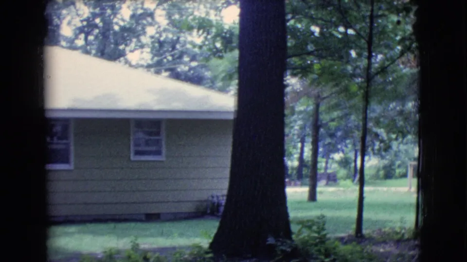 1961 Clarkdale, Arizona home-movie frame showing a tan house, pale sloped roof, windows, lawn, and tree trunks in the foreground.