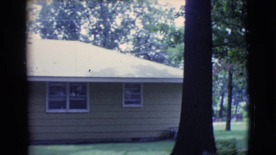 1961 Clarkdale, Arizona frame showing the side of a tan residential house with windows, a pale roof, tree trunks, and lawn.