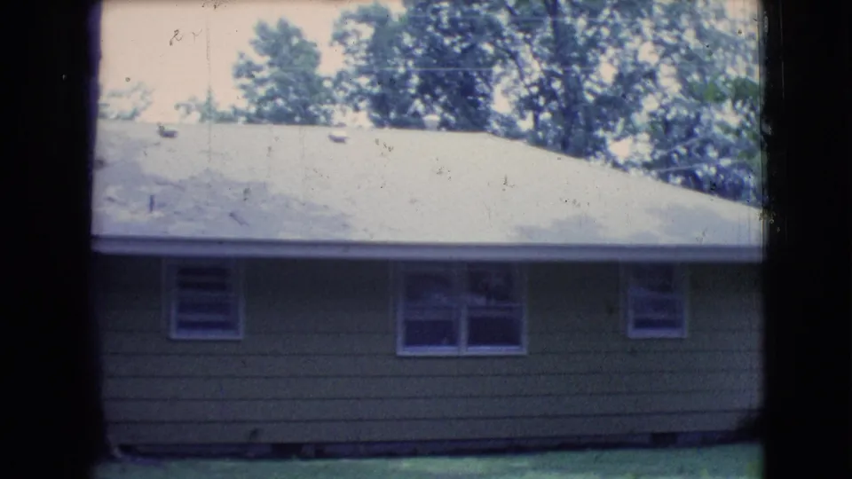 1961 Clarkdale, Arizona frame with a closer view of a tan house exterior, broad pale roof, small windows, grass, and trees.