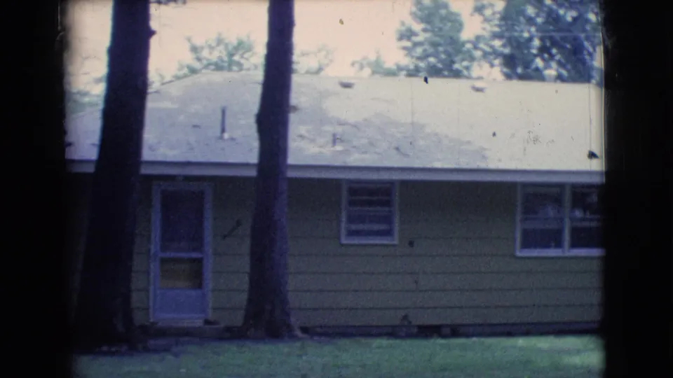1961 Clarkdale, Arizona frame showing a tan house exterior with a door, windows, pale roof, mature tree trunks, and grass.