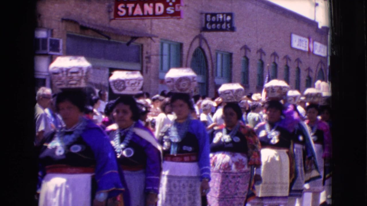 Women in colorful parade attire carry patterned baskets on their heads past storefront signs.