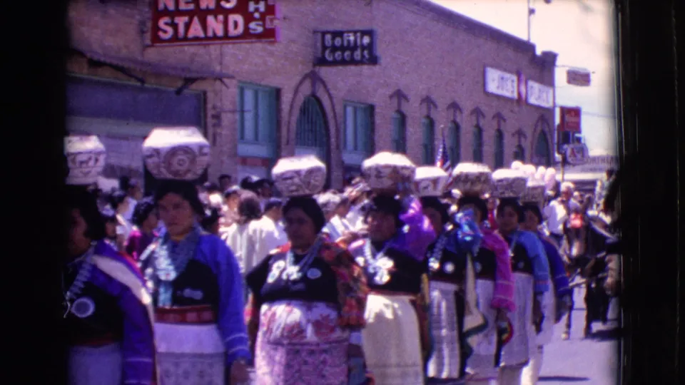 Women in coordinated garments continue down the Clarkdale street during the 1961 parade, with storefront signage and spectators behind them.